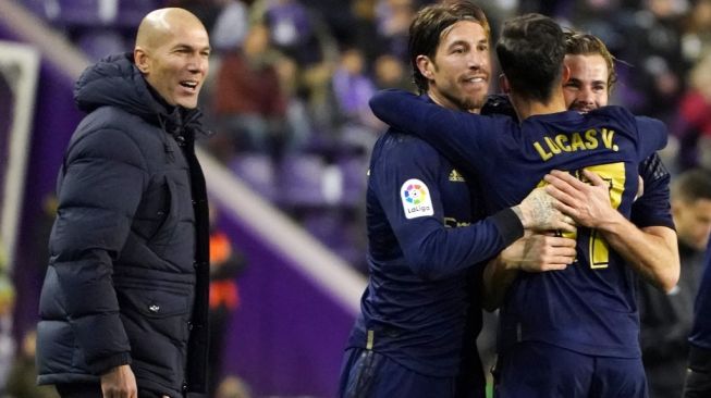 Pelatih Real Madrid, Zinedine Zidane (kiri) menyaksikan dari pinggir lapangan saat para pemain Real Madrid merayakan gol ke gawang Real Valladolid pada laga lanjutan Liga Spanyol 2019/2020. [CESAR MANSO / AFP]